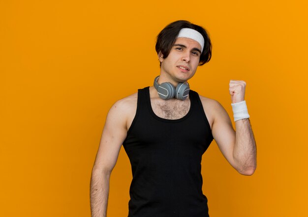 Young sporty man wearing sportswear and headband with headphones around neck with confident expression pointing back 
