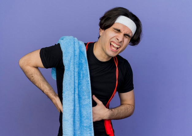 Молодой спортивный мужчина в спортивной одежде и повязке на голову со скакалкой на шее и полотенцем на плече выглядит неприветливо и страдает от боли в животе