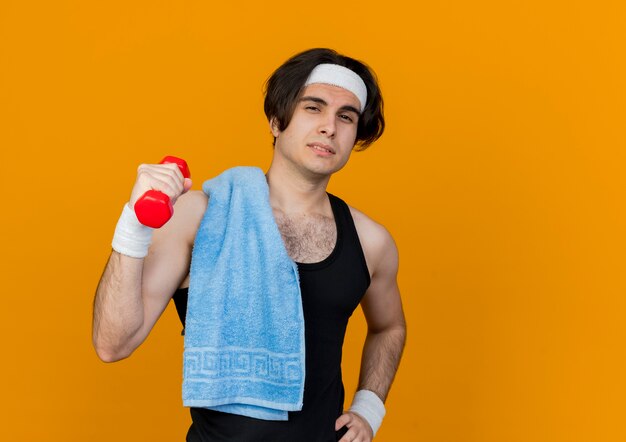 Young sporty man wearing sportswear and headband with towel on shoulder holding dumbbell looking confident 
