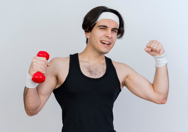 Young sporty man wearing sportswear and headband working out with dumbbell raising fist showing biceps 