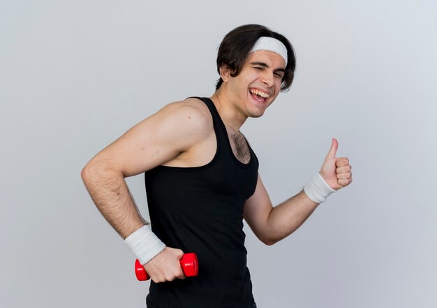 운동복과 머리띠를 착용하는 스포티 한 젊은이가 행복하고 긍정적 인 미소를 보여주는 흰 벽 위에 서있는 엄지 손가락으로 운동