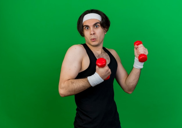young sporty man wearing sportswear and headband working out with dumbbells looking confused 