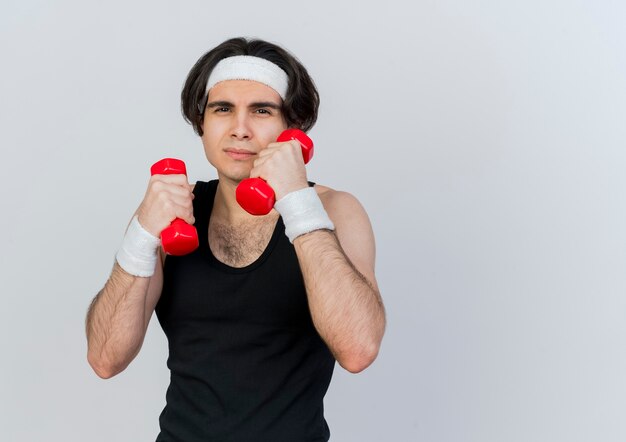 Young sporty man wearing sportswear and headband working out with dumbbells looking at front with serious face standing over white wall