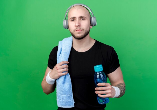 молодой спортивный мужчина в браслете и наушниках держит бутылку с водой с полотенцем на плече