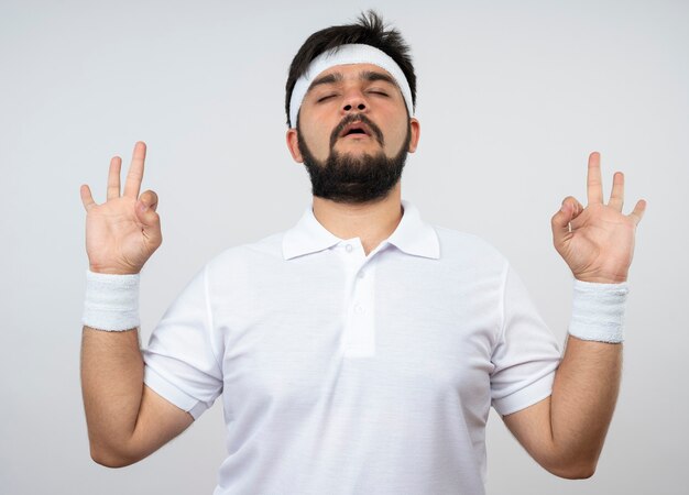 Young sporty man with closed eyes wearing headband and wristband showing meditation gesture isolated on white wall