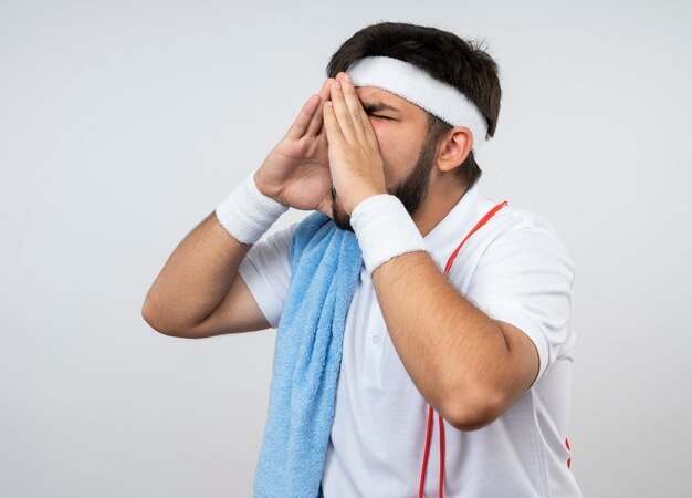 Young sporty man with closed eyes wearing headband and wristband with towel and jump rope on shoulder calling someone isolated on white wall