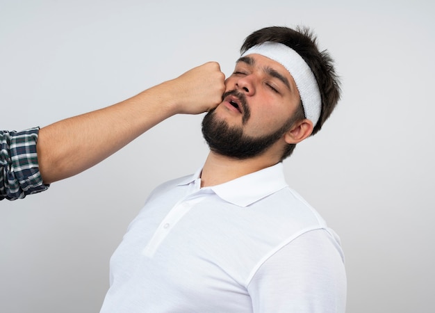 Young sporty man with closed wearing headband and wristband beating by someone isolated on white wall