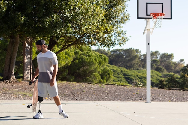 Young sporty man with leg prosthesis leaving basketball court after skateboarding there. Confident bearded guy with disability holding skateboard while doing sports outdoors. Amputee sport concept