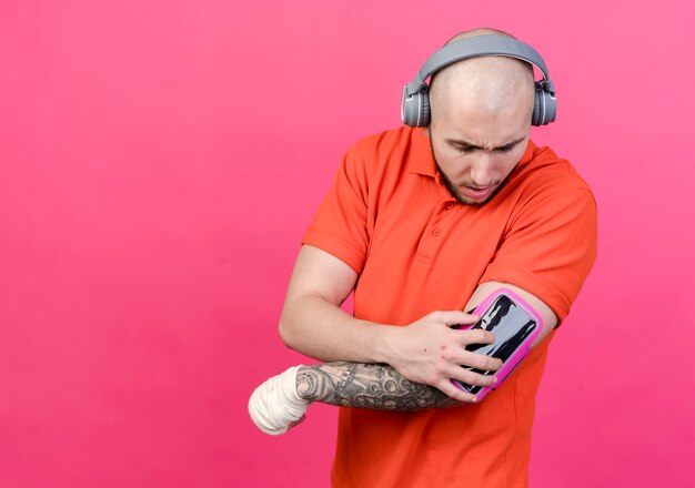손목 붕대 헤드폰을 착용하고 분홍색 벽에 고립 된 그의 팔에 전화 팔 밴드를보고 스포티 한 젊은이