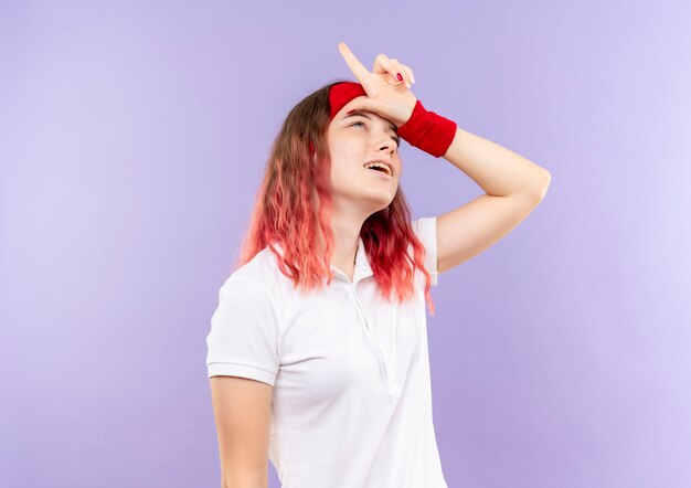 Young sporty woman doing loser sign over her head smiling standing over purple wall