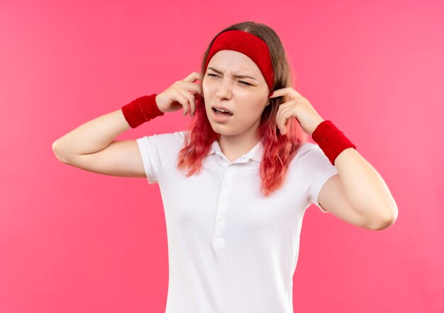 Young sporty woman in headband closing ears with fingers with annoyed expression standing over pink wall