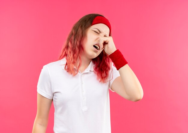 Young sporty woman in headband closing her nose with fingers suffering from stench standing over pink wall