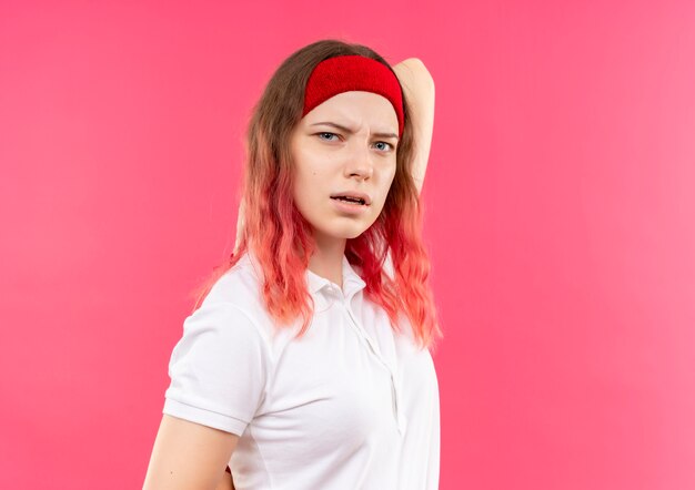 Young sporty woman in headband confused and uncertain standing over pink wall