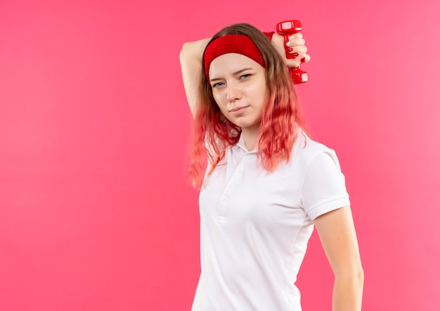 Young sporty woman in headband doing exercises with dumbbell looking confident standing over pink wall