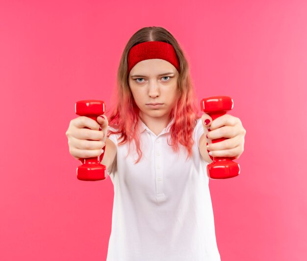 Young sporty woman in headband doing exercises with dumbbells with serious confident expression standing over pink wall