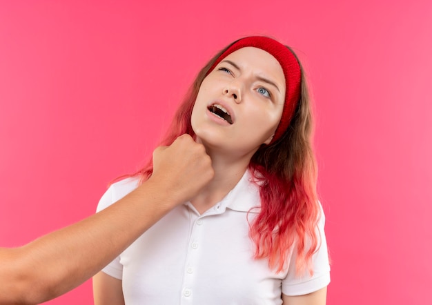 Free photo young sporty woman in headband getting puch in her jaw with fist standing over pink wall