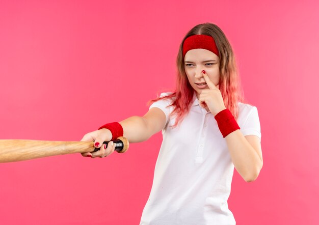 Young sporty woman in headband going to play baseball holding a bat, looking confident standing over pink wall