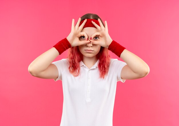 Young sporty woman in headband holding fingers in binoculars gesture with fingers through fingers standing over pink wall