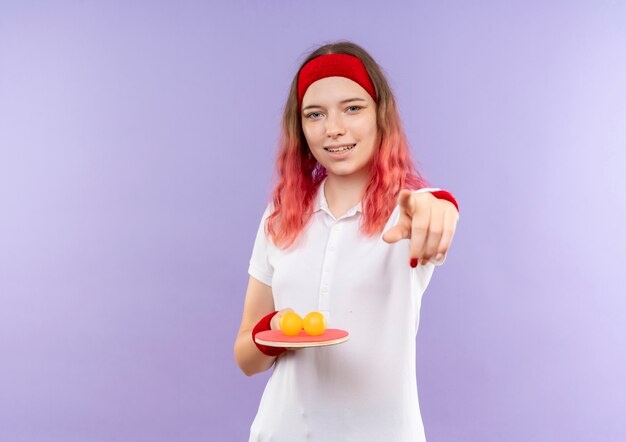 Young sporty woman in headband holding racket for table tennis and balls pointing with finger to camera smiling standing over purple wall