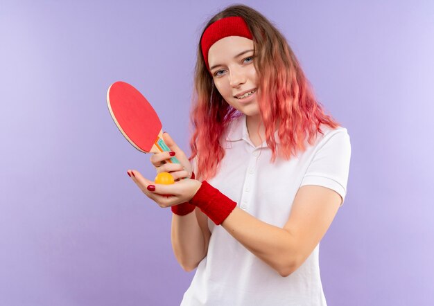Молодая спортивная женщина в повязке на голову, держащая ракетку для настольного тенниса и мячи с улыбкой на лице, стоящая над фиолетовой стеной