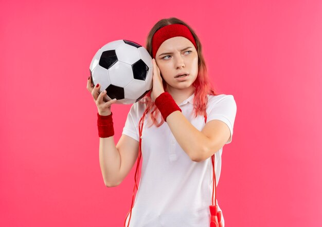 Young sporty woman in headband holding soccer ball looking aside worried standing over pink wall