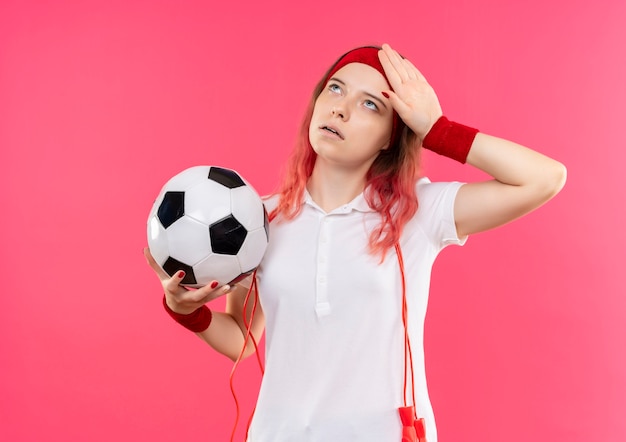 Young sporty woman in headband holding soccer ball looking tired and exhausted standing over pink wall