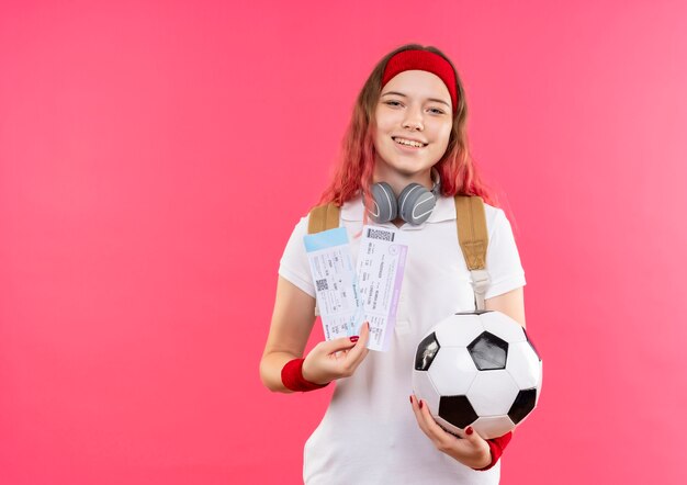 Young sporty woman in headband holding two air tickets and soccer ball smiling cheerfully standing over pink wall