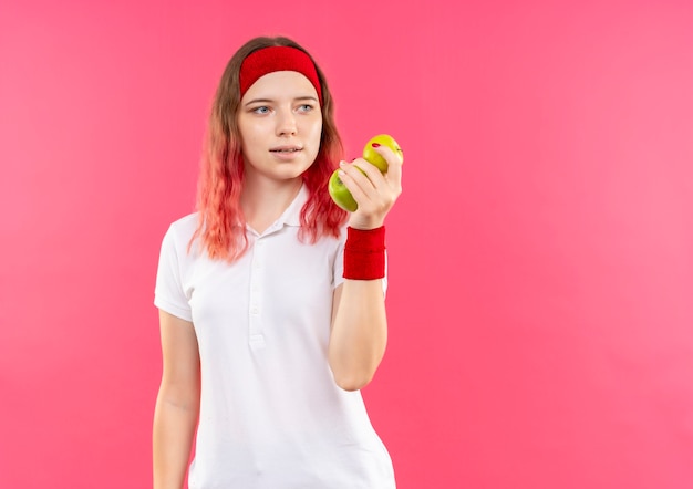 Young sporty woman in headband holding two apples looking aside positive and happy standing over pink wall