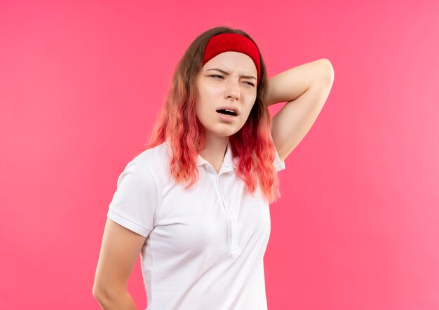 Young sporty woman in headband looking aside confused touching her neck having pain standing over pink wall