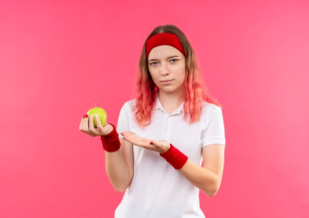분홍색 벽 위에 자신감 서 찾고 그녀의 손의 팔으로 녹색 사과를 제시하는 머리띠에 젊은 스포티 한 여자