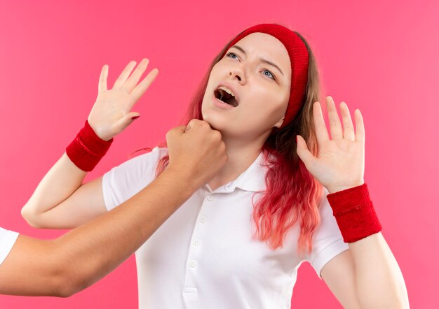 Молодая спортивная женщина в повязке на голову, подняв кулаки в знак капитуляции, рвет ей в лицо, когда кто-то кулак стоит над розовой стеной