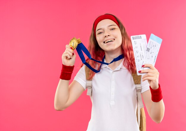 Young sporty woman in headband showing her gold medal holding two air tickets smiling with happy face standing over pink wall