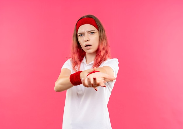 Free photo young sporty woman in headband surprised and amazed stretching her arm standing over pink wall