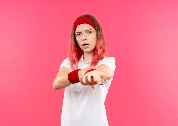 Young sporty woman in headband surprised and amazed stretching her arm standing over pink wall