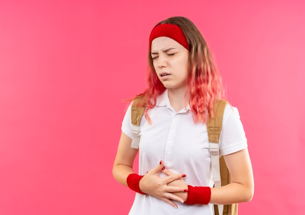 Young sporty woman in headband touching her belly looking unwell feeling pain standing over pink wall