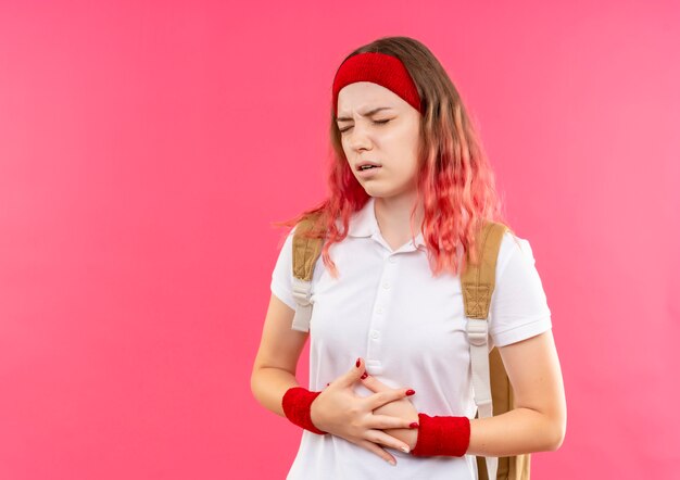 Молодая спортивная женщина в повязке на голову, касаясь ее живота, выглядит нездоровой, чувствуя боль, стоя над розовой стеной