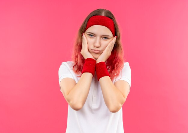 Молодая спортивная женщина в повязке на голову, касаясь ее лица руками, выглядит усталой и скучающей, стоя у розовой стены
