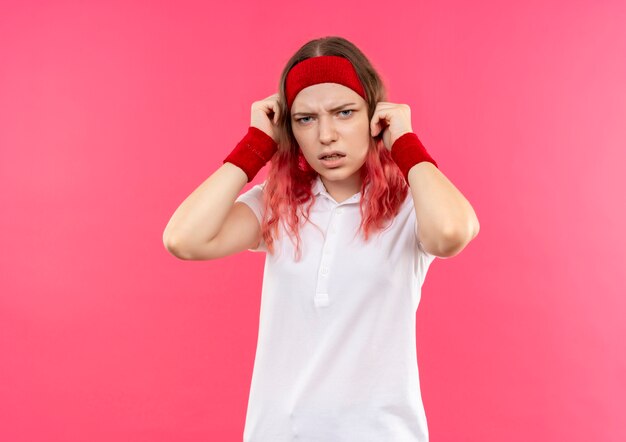 Young sporty woman in headband with annoyed expression standing over pink wall