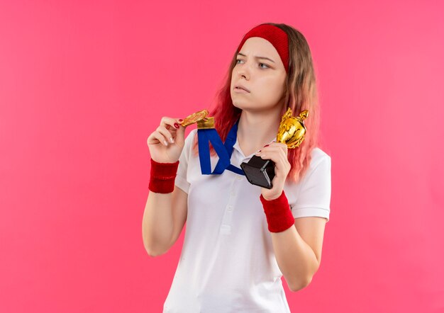 Young sporty woman in headband with gold medal around her neck holding trophy looking aside with serious expression standing over pink wall