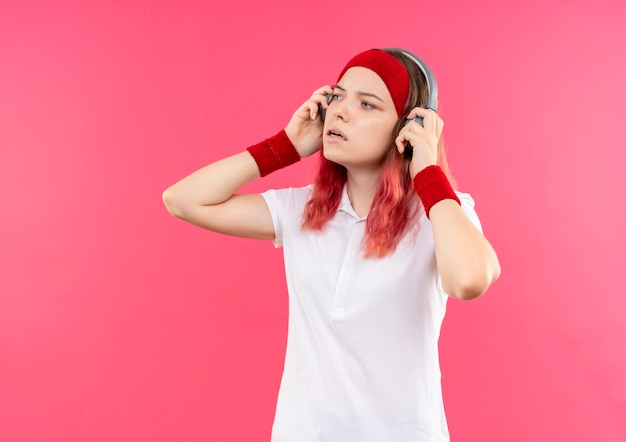 Free photo young sporty woman in headband with headphones listening to music looking aside standing over pink wall