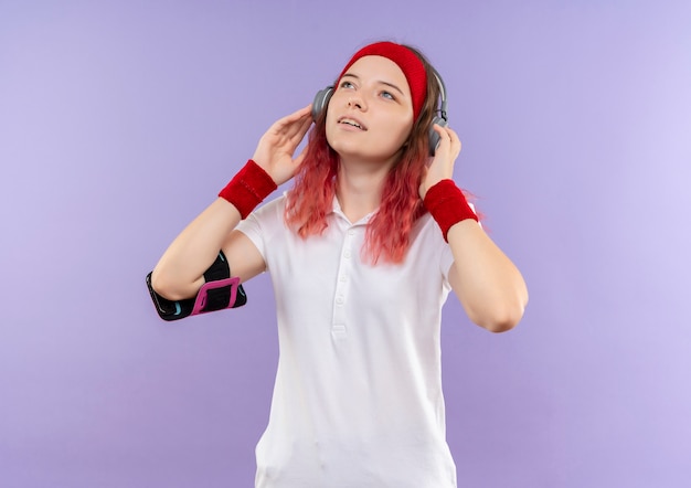 彼女のお気に入りの音楽を楽しんで見上げるヘッドフォンでヘッドバンドの若いスポーティな女性、紫色の壁の上に立っているスマートフォンの腕章でトレーニング