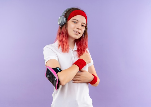 Young sporty woman in headband with headphones smiling with smartphone armband standing over purple wall