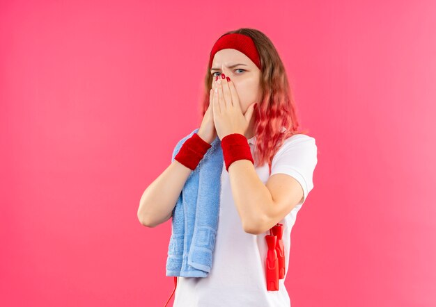 Young sporty woman in headband with towel on shoulder shocked, covering mouth with hands standing over pink wall