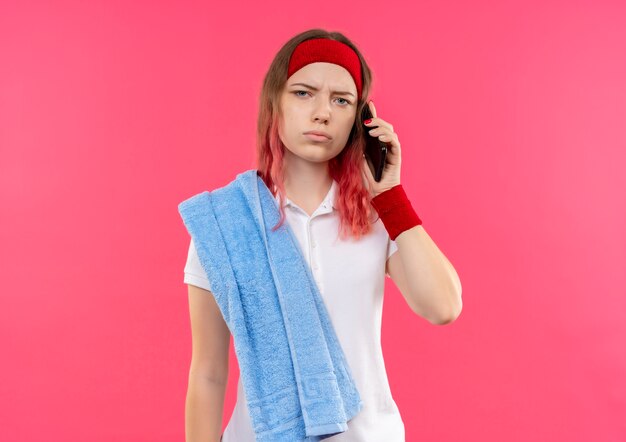 Молодая спортивная женщина в повязке на голову с полотенцем на плече разговаривает по телефону monile, выглядит смущенной и очень взволнованной, стоя у розовой стены