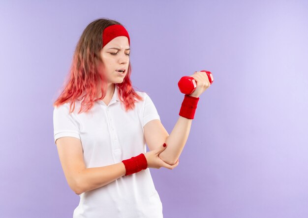 Молодая спортивная женщина, держащая одну гантель, делает упражнения, глядя на локоть, чувствуя боль, стоя над фиолетовой стеной