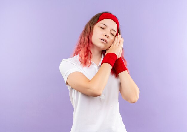 Young sporty woman holding palms together leaning head on palms looking tired wants to sleep standing over purple wall