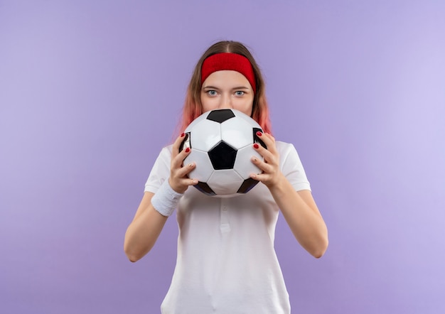 Молодая спортивная женщина держит футбольный мяч, пряча лицо за мячом, стоящим над фиолетовой стеной