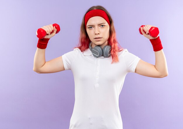 Young sporty woman holding two dumbbells doing exercises with serious face standing over purple wall