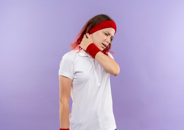 Young sporty woman looking unwell touching her neck feeling pain standing over purple wall