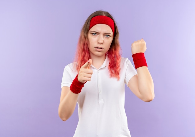 Giovane donna sportiva che indica con il dito indice al pugno di serraggio della fotocamera con la faccia seria in piedi sopra la parete viola